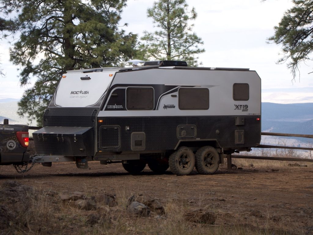 An MDC USA XT19 Off-Grid Extreme trailer parked on rugged terrain near a scenic mountain overlook, surrounded by tall pine trees. The black and white off-road travel trailer is attached to a towing vehicle, showcasing its durable build and all-terrain capabilities for off-grid adventures.